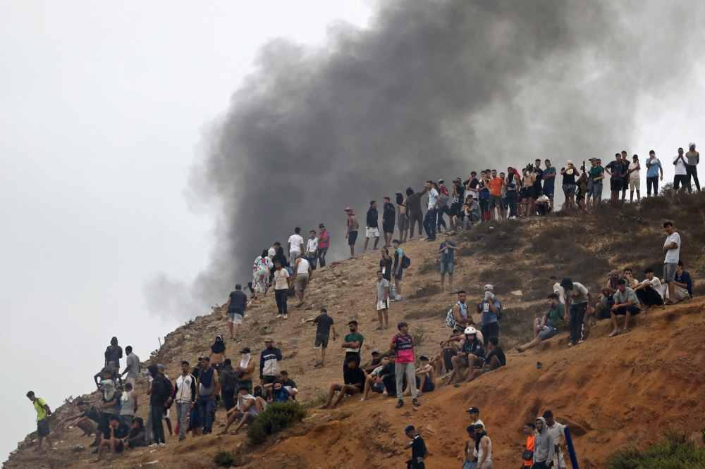 People gather on a hilltop as smoke billows from the site of clashes near Fnideq on the Morocco-Spain border on July 31, 2026. — AFP pic